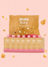 A bag of Shake Baby grain-flavored protein shakes with six individual squeeze bottles lined up in front, set against a soft pink background with scattered popcorn pieces.