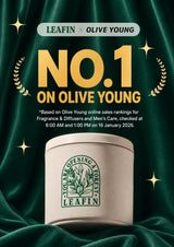 A promotional graphic for a LEAFIN x OLIVE YOUNG fragrance product. A cream-colored cylindrical tin with a green LEAFIN logo sits on draped dark green fabric, surrounded by gold starbursts and laurel wreaths.