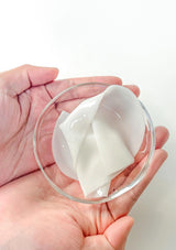Hand holding a small glass dish with a white gel-like substance against a white background