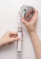 A person's hands are shown disassembling a clear shower head filter cartridge, holding the transparent shower head and white filter core against a plain white background.
