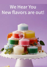 A pyramid arrangement of colorful wax melts or scented bars in small clear jars on a white pedestal, with decorative flowers and citrus slices, against a lavender background. White text at the top reads, 'We Hear You New flavors are out!'.
